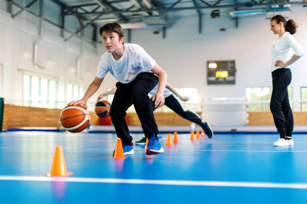 Students and their coach are in school gym and have training class. Teacher give instructions to pupils and watch them with attention. Boys are running in precise order between the cones with basketball. They are wearing sports uniform and warming up.